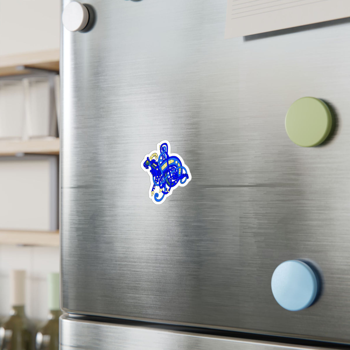 A small blue illustrated sticker of a Viking Age Jelling art style goat with a white border is attached to a stainless steel refrigerator door, alongside other colorful fridge magnets in the background.