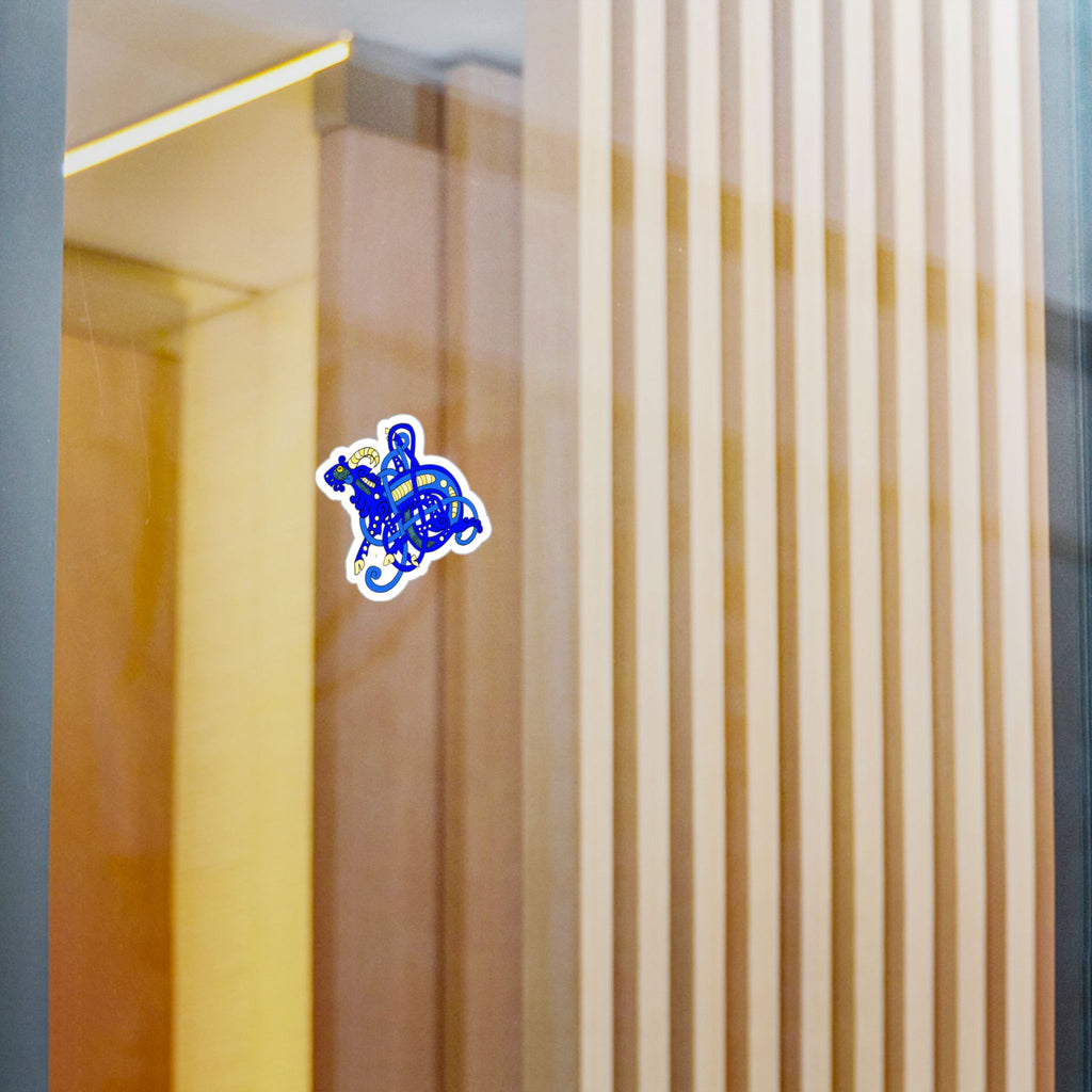 A small die-cut sticker featuring a blue Viking Age Goat Jelling Art style applied to a glass door or window with a warm-toned interior visible behind it.