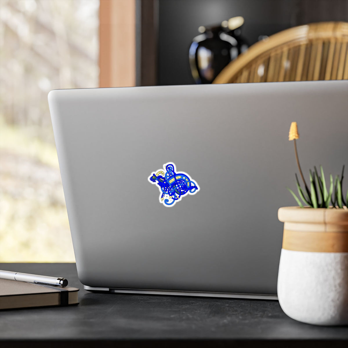 Laptop with a colorful Viking Age goat jelling art style sticker on a desk with a plant and pen in the background